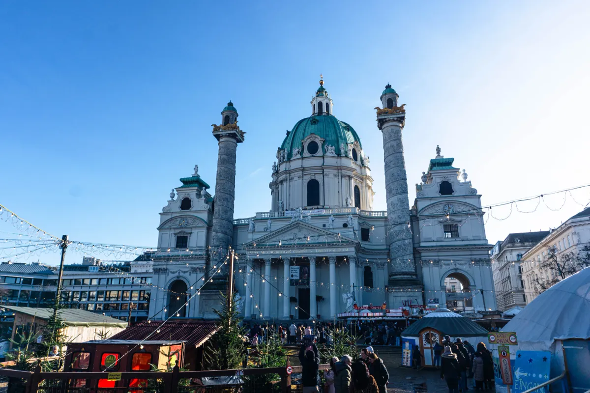A baroque church with a green copper dome and two tall decorated columns, with a Christmas market stall in front.