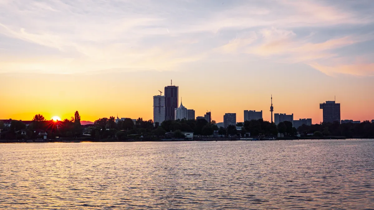 The sun sets behind Vienna's skyline across a lake, with silhouetted towers and a TV tower under an orange sky.