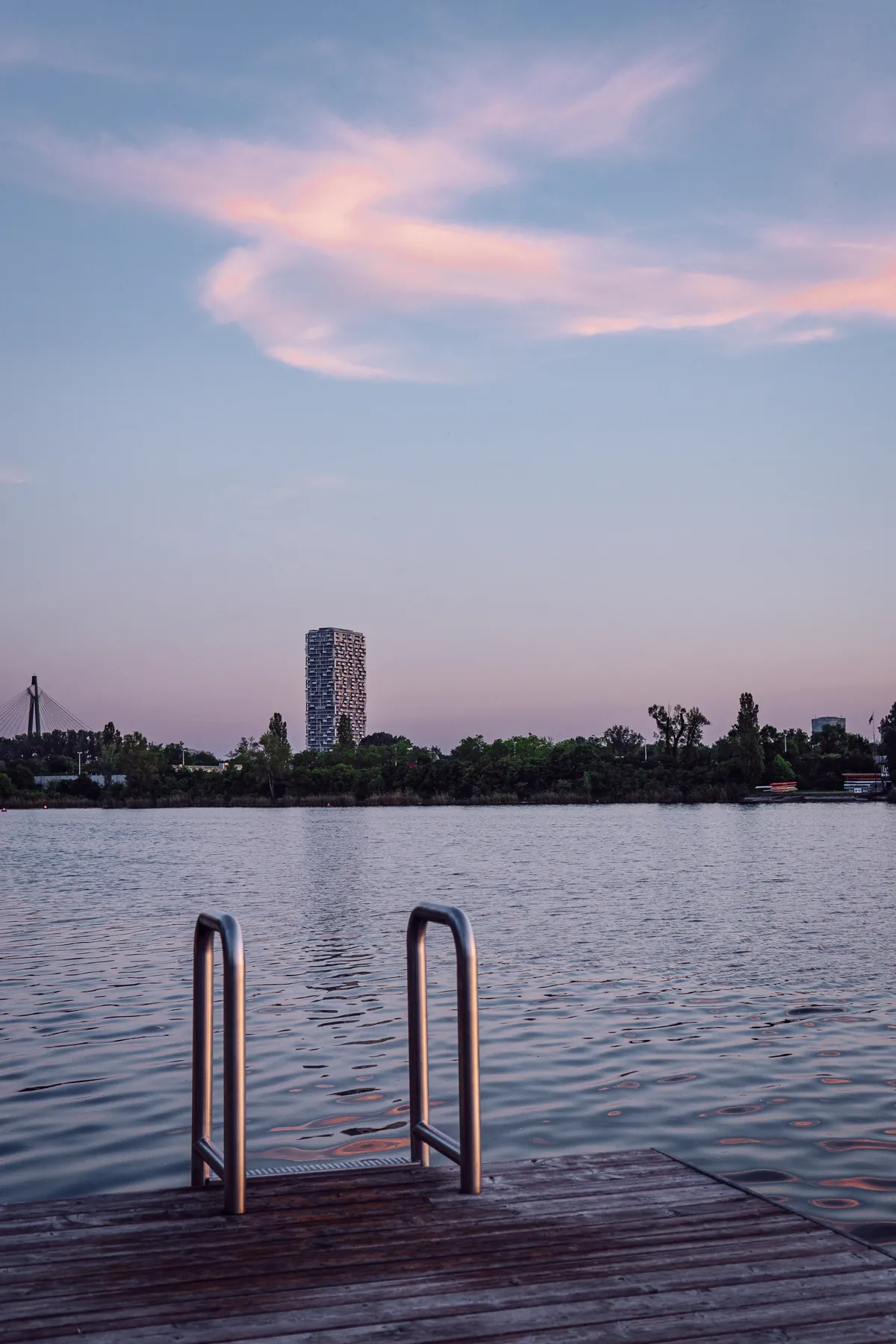 A wooden jetty with steel ladder rails over a calm lake at dusk, with a tower block and treeline beyond.
