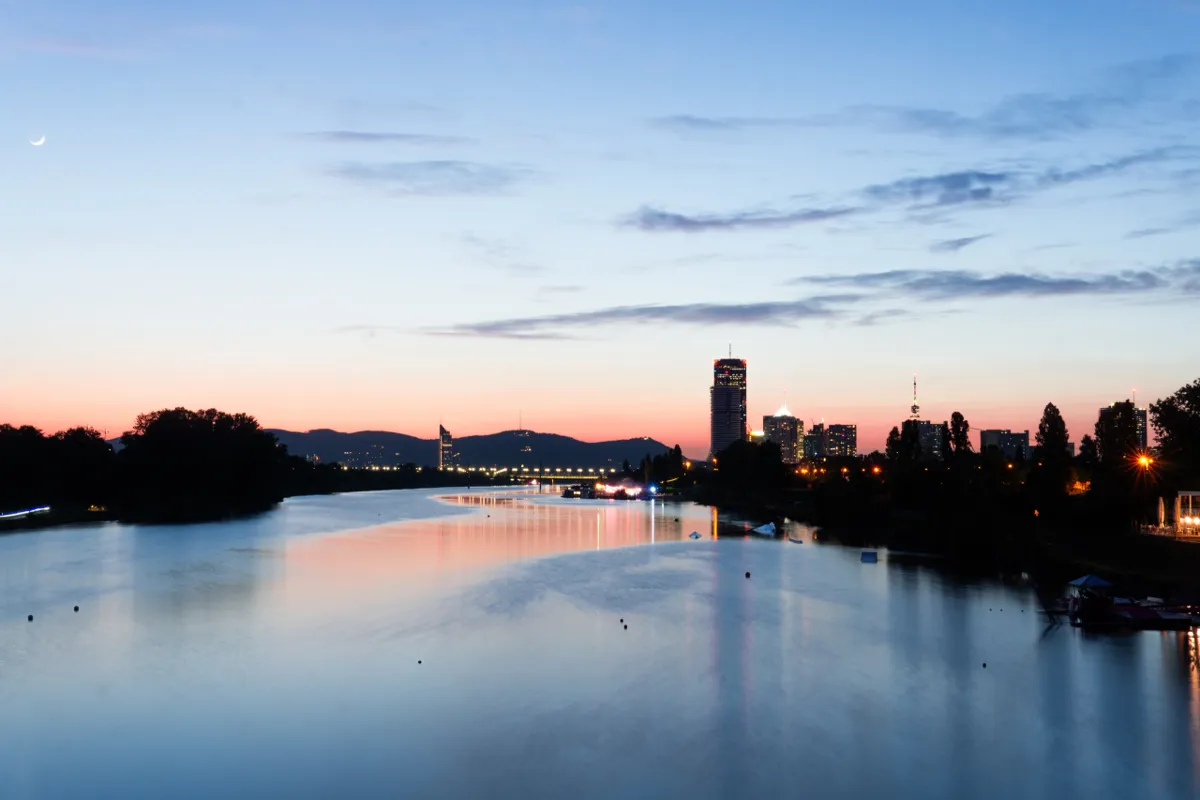 A river in Vienna at dusk reflects a pink and blue sky, with silhouetted trees and tall buildings beyond.