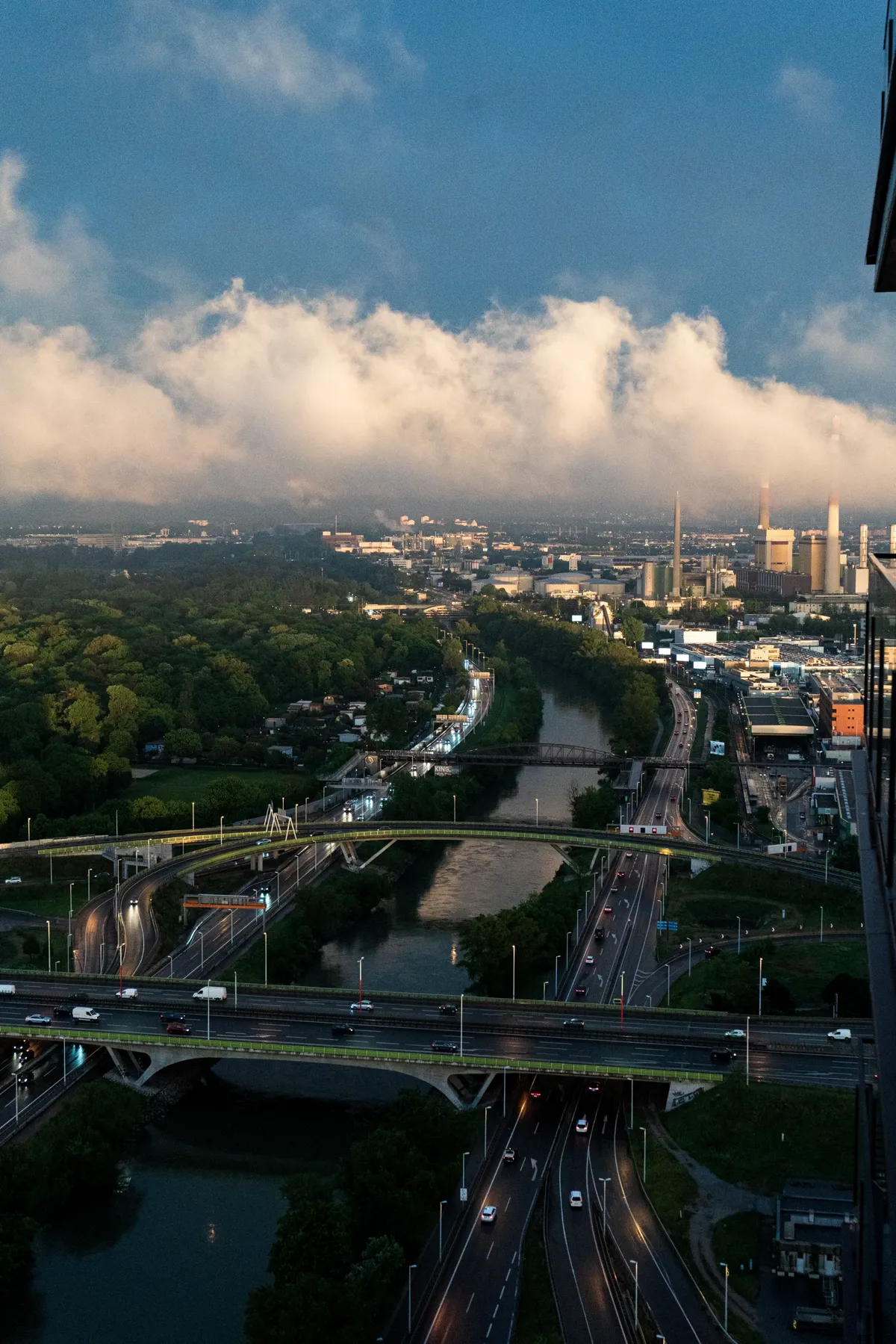 Bridges and motorways cross a river in Vienna, with green woodland and a glowing bank of clouds above at dusk.